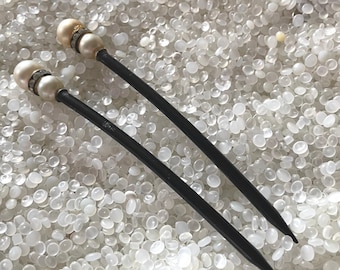 May include: Two black hair sticks with pearl and rhinestone accents. The top of each stick features a faux pearl and a band of small rhinestones. The sticks are long and slender, with a pointed end.