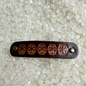 May include: A dark brown leather bracelet with five embossed flower designs. The bracelet is rectangular with a metal stud at each end. The flowers are a lighter brown colour.