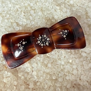 May include: A tortoiseshell-patterned hair clip in a bow shape, adorned with sparkling rhinestones. The clip features a dark brown and amber color scheme, with the rhinestones clustered in decorative patterns.