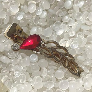 May include: A gold-tone metal hair clip featuring an openwork braided design. The clip is accented with a single teardrop-shaped red gemstone and a small, round, clear faceted stone positioned near the hinge mechanism of the hair accessory.