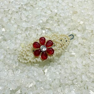 May include: A decorative hair clip featuring a red flower with a clear crystal center. The flower is set on a base of faux pearls. The clip has a silver-colored metal clasp.