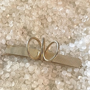May include: A gold-toned metal barrette with a mesh-like texture and a decorative looped design. The barrette is a hair accessory, likely for securing hair in place. The background is a light-colored, textured surface.