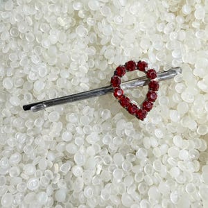 May include: A silver-toned hair barrette featuring a teardrop-shaped design adorned with numerous small, round, red gemstones. The barrette is set against a backdrop of small, white, rounded objects.