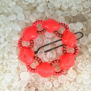 May include: A bright red vintage hair barrette featuring a seashell design. The barrette is made of plastic and has a black metal clasp. The seashell design is arranged in a circular pattern.