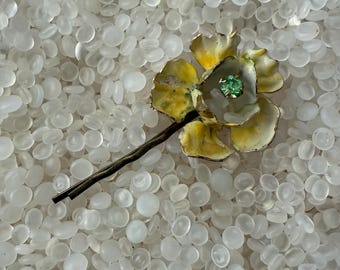 May include: A vintage floral hair clip featuring a yellow and white flower with a green center stone. The flower is attached to a dark brown metal stem and hair clip. The clip is set against a background of small, white, oval-shaped beads.