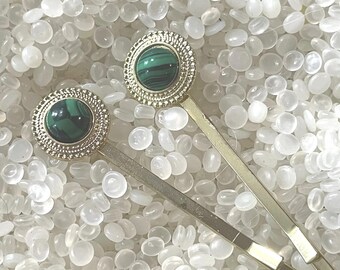 May include: Two gold-toned bobby pins with round, decorative tops. Each top features a dark green malachite stone surrounded by a textured, gold-colored frame. The bobby pins are set against a background of small, white, rounded objects.