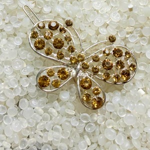May include: A silver-toned butterfly hair clip with amber-colored rhinestones. The butterfly design features openwork wings and a central body, with the rhinestones adding sparkle. The clip is set against a white background.