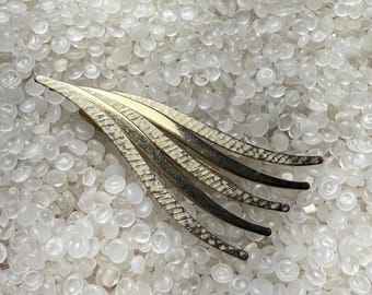 May include: A vintage gold-tone hair clip with a leaf-like design. The clip features multiple slender, curved elements with textured detailing, transitioning from gold to a darker hue at the tips. The clip is set against a backdrop of small, translucent white beads.
