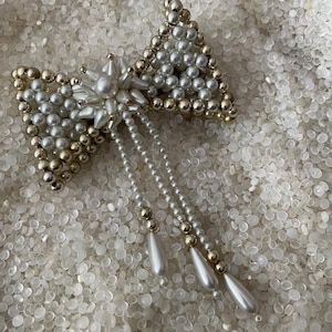 May include: A large, white and gold pearl bow with a dangling pearl tassel. The bow is made of many small pearls and is a statement piece.