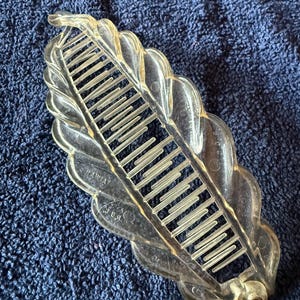 May include: A clear, plastic hair comb in a leaf shape, with a series of teeth for holding hair in place. The comb has a decorative, scalloped edge and a small, round peg at the base. The comb is lying on a blue, textured surface.