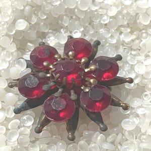 May include: Vintage brooch featuring a flower design with faceted, deep red glass stones. The brooch has a bronze-toned metal setting with pointed accents between the red stones. A classic piece of jewelry.