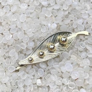 May include: A gold-toned leaf-shaped hair clip with four spherical, silver-colored accents. The clip has a textured surface and is set against a background of small, white, oval-shaped beads. The clip is likely made of metal.