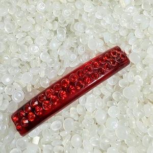May include: A rectangular, red bar with embedded red rhinestones. The bar is set against a background of small, white, translucent, oval-shaped beads. The rhinestones are arranged in rows along the bar's surface.