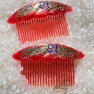 May include: Two red hair combs with a floral design. The combs have a scalloped top and long, thin teeth. The floral design features purple roses, green leaves, and gold accents.