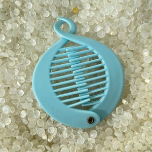 May include: A light blue vintage hair comb with a unique design. The comb features a curved handle and a fan-shaped section with horizontal teeth. The comb is made of plastic and has a small, round, dark-colored rivet. The comb is set against a background of small, white, rounded objects.