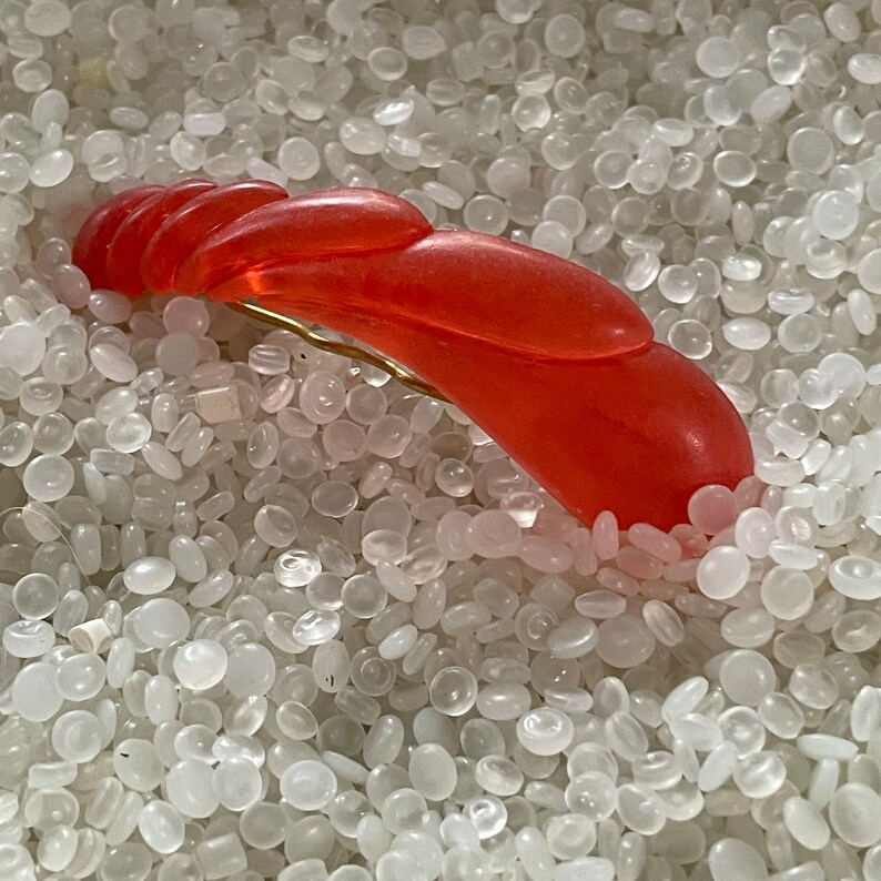 May include: A vibrant orange hair clip with a curved, layered design. The clip features a gold-colored metal clasp. The accessory is set against a background of small, white, rounded objects.