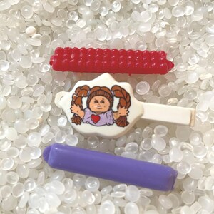 May include: Three hair accessories including a red rectangular clip with a textured surface, a white hair clip featuring a graphic illustration of a doll with brown pigtails and a red heart on a purple shirt, and a smooth purple rectangular hair clip.