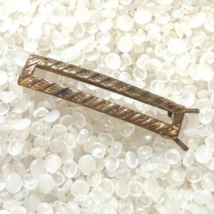 May include: A rectangular gold-tone metal hair clip featuring an open center design and a textured, diagonal striped pattern along the frame. The accessory has a hinged mechanism on one end and a smooth, metallic finish throughout the structure.