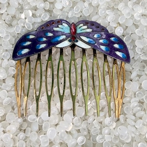 May include: A hair comb featuring a decorative butterfly top with purple, blue, and white enamel detailing. The butterfly has a small red accent at the center. The comb has nine gold-toned metal teeth extending downward from the butterfly base.