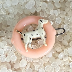 May include: A vintage brooch featuring a white Dalmatian dog with black spots, standing inside a light pink ring. The brooch has a silver-tone pin on the back. The background is filled with small, translucent, oval-shaped beads.