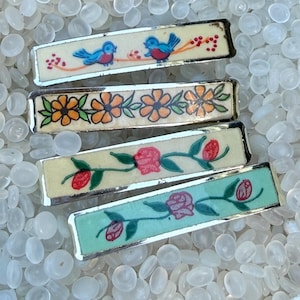 May include: Four rectangular hair clips with silver-tone metal frames. The top clip features blue birds on a cream background. The second has orange flowers with green leaves. The third shows red flowers on cream. The bottom has red roses on mint green.
