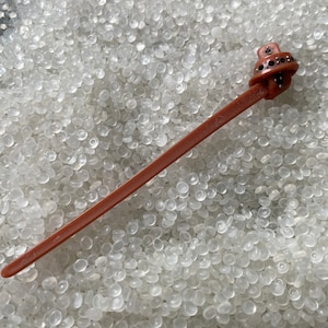 May include: A brown plastic cable tie with a black and silver decorative end. The cable tie is lying on a white surface.