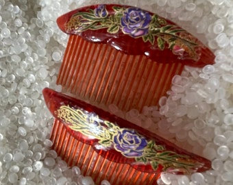 May include: Two vintage red hair combs with floral designs. The combs feature a curved top with a painted design of a purple rose and gold accents. The teeth of the combs are a lighter shade of red. The combs are resting on a white surface.