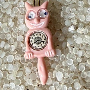 May include: A pink cat-shaped clock with a smiling face, large eyes, and a tail. The clock face has black numbers and hands. The cat is wearing glasses. The clock is set to 2:50.