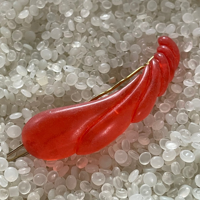 May include: A vintage, coral-colored hair barrette with a swirling, textured design. The barrette is made of a translucent material and features a gold-tone metal pin for securing it in the hair. A unique hair accessory.