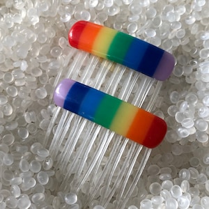 May include: Two rainbow hair clips with a purple, blue, green, yellow, and orange color scheme. The clips are on a white background.