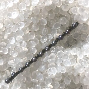 May include: A dark gray beaded bracelet with small, round, dark gray pearls and smaller, bronze-colored beads. The bracelet is strung on a dark cord and is displayed against a white, textured background.
