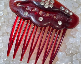 May include: A vintage hair comb with a dark red-brown, curved top and long, thin teeth. The top is adorned with clear, sparkling rhinestones in a floral and dot pattern. The comb is a classic hair accessory.