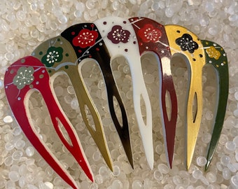 May include: A colorful hair comb with five prongs, each painted with a different floral design. The prongs are red, gold, black, white, and green.