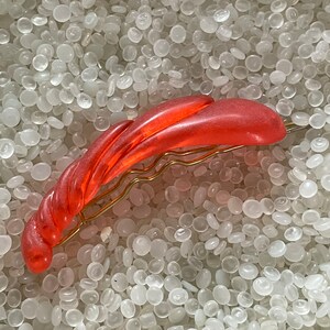May include: A coral-colored hair barrette with a twisted, textured design. The barrette is made of a translucent material and features a gold-toned metal clasp. This hair accessory is suitable for various hairstyles.