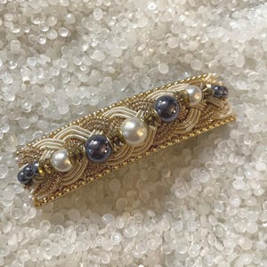 May include: A rectangular hair barrette with a gold-colored frame, adorned with woven gold and cream-colored threads, and embellished with white and dark gray pearls. The barrette is a decorative hair accessory.
