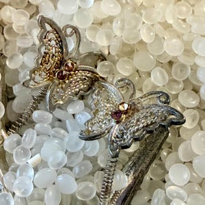 May include: A silver-toned hair accessory featuring two butterfly-shaped ornaments with intricate filigree wings and small, round, reddish-brown stones set in the center of each butterfly body, attached to a flexible metal coil spring base.
