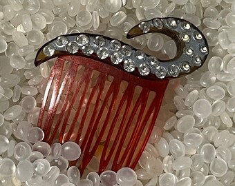 May include: A vintage hair comb with a red base and a decorative top. The top features a curved, dark-colored design adorned with numerous clear rhinestones. The comb's teeth are a deep red color.