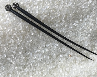 May include: Two black hair sticks with decorative, dark-colored, spherical embellishments at the top. The hair accessories are set against a background of small, white, rounded objects.