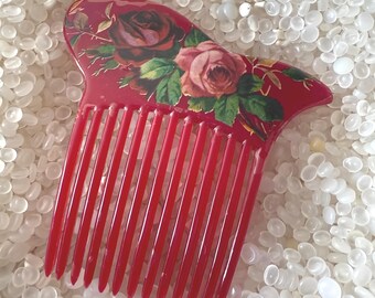 May include: A red hair comb with a floral design featuring roses in shades of red and pink. The comb has a curved top and long, evenly spaced teeth. The floral design is on the top of the comb.