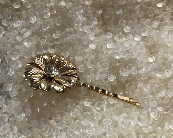 May include: A gold-colored metal hair barrette shaped like a flower with a clear rhinestone center. The barrette has a simple, elegant design, perfect for adding a touch of sparkle to any hairstyle.