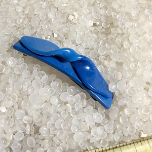 May include: A bright blue hair clip with a flowing, abstract shape. The clip, made from a shiny material, is set against a backdrop of small, white, translucent beads. The design includes a central, rounded feature.