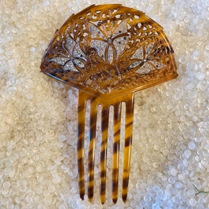 May include: A brown tortoiseshell hair comb with a decorative fan-shaped top. The top is intricately carved with a floral design and features small, clear rhinestones.