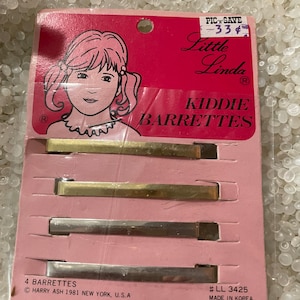 May include: A pink and white packaging card with a cartoon girl on the front. The card says "Little Linda Kiddie Barrettes" and "4 Barrettes". The back of the card says "Harry Ash 1981 New York, U.S.A" and "#LL 3425 Made in Korea".