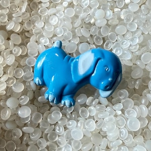 May include: A bright blue dog figurine with white accents on its ears and snout. The dog is lying down, with its tail raised. The figurine is set against a background of small, white, rounded objects.