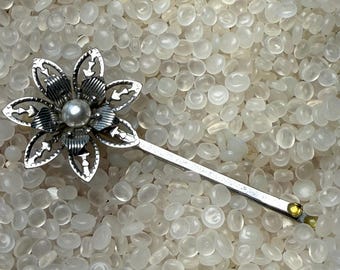 May include: A silver-toned hair barrette with a flower design. The barrette features a central pearl-like bead and openwork petals. The stem of the barrette is straight and ends with a small yellow accent. The barrette is set against a background of small, translucent, white beads.