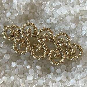 May include: A gold-toned hair barrette with a repeating floral design. Each flower is formed by a twisted rope-like texture, with a small ball in the center. The barrette is set against a background of small, white, oval-shaped objects.