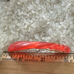 May include: A coral-colored vintage hair clip with a twisted design. The clip is made of plastic and has a gold-tone metal clasp. The ruler shows the clip is approximately 3 inches long.