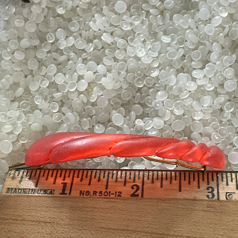 May include: A coral-colored vintage hair clip with a twisted design. The clip is made of plastic and has a gold-tone metal clasp. The ruler shows the clip is approximately 3 inches long.