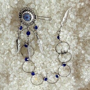 May include: A silver-toned hair clip with a circular design, adorned with blue and white accents, dangling chains, and small silver feathers. The clip features a series of connected rings and blue gemstone accents, creating a bohemian aesthetic.