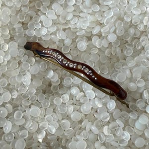 May include: A wavy hair barrette featuring a dark brown tortoiseshell-style finish with a central row of small, clear, round rhinestones embedded along the length of the piece, attached to a thin, gold-toned metal spring-loaded clasp mechanism.
